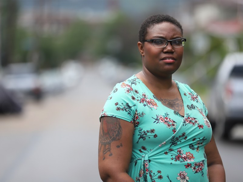 Tanisha Bowman, wearing a blue floral dress and glasses, stands outside posing for a photo.